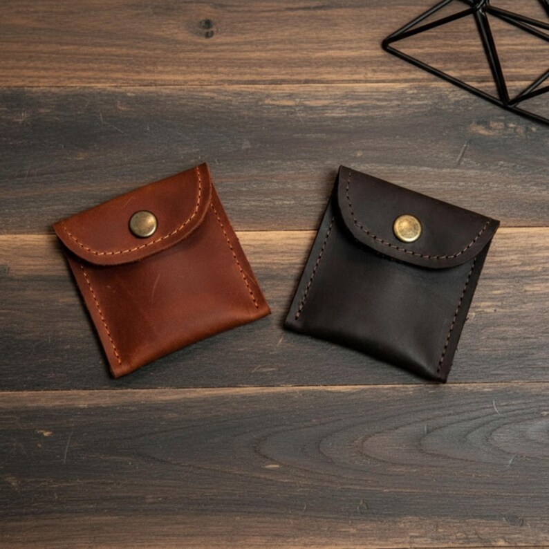 May include: Two small, square leather pouches in shades of brown and dark brown. Each pouch has a flap closure secured by a brass button. The pouches are stitched with contrasting thread, suggesting handmade craftsmanship.