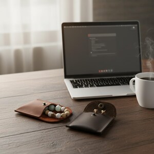 May include: A brown leather pouch with a snap closure, open to reveal a beaded bracelet with cream, green, and tan beads. A second, smaller dark brown leather pouch is open, displaying small earrings. A laptop and coffee cup are in the background.