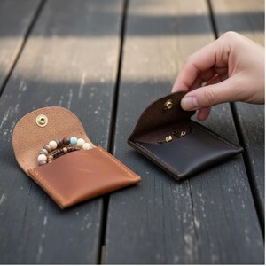 May include: Two small, open leather pouches. One is light brown and contains beaded bracelets with tan, brown, and turquoise beads. The other is dark brown and holds small gold earrings. Both pouches have a snap closure.