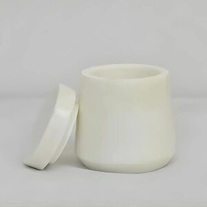 Pode incluir: Um recipiente cilíndrico liso de cor branco-sujo com uma base ligeiramente cónica e uma tampa circular a condizer colocada na vertical ao lado. O objeto apresenta um acabamento sólido e opaco com um design simples e uma abertura larga.