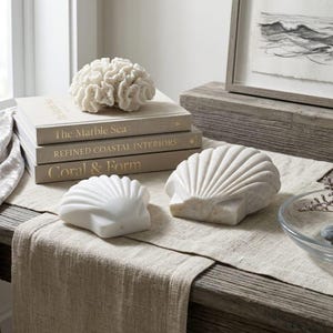 May include: Two white marble seashell sculptures placed on a beige linen runner. Behind them, a stack of three books titled The Marble Sea, Refined Coastal Interiors, and Coral & Form, topped with a white coral decorative piece.
