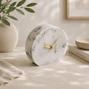Puede incluir: Un reloj de escritorio circular hecho de mármol blanco con vetas grises. La esfera del reloj presenta una superficie de piedra lisa y pulida con dos manecillas de metal fino de color dorado situadas en el centro para indicar la hora sin marcas.