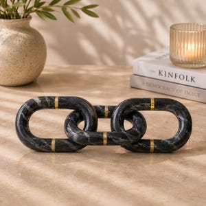 Puede incluir: Una escultura decorativa de cadena hecha de mármol negro con detalles dorados. La escultura está compuesta por tres eslabones ovalados entrelazados. Los libros The Kinfolk y A Democracy of Image están en el fondo.