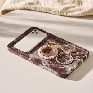 Puede incluir: Una bandeja rectangular de mármol color granate oscuro con vetas blancas. Sobre la bandeja hay un teléfono inteligente blanco, una pulsera de cuentas esféricas de color rosa claro, una pulsera de cadena dorada y un pendiente de aro fino.