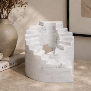 May include: A white marble sculpture featuring a spiral staircase design with a cylindrical base. The stone displays natural grey veining throughout the structure, which consists of stacked rectangular blocks forming a continuous winding path.