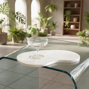 Puede incluir: Una copa transparente llena de líquido descansa sobre una bandeja de mármol blanco en forma de riñón. La bandeja está sobre una mesa de cristal con un borde verde azulado. El fondo presenta plantas de interior y ventanas arqueadas.