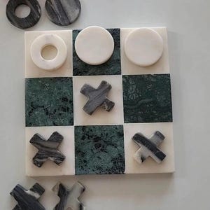 Puede incluir: Un tablero de juego de tres en raya cuadrado hecho de baldosas de mármol blancas y verde oscuro alternas. El conjunto incluye varias piezas circulares blancas, algunas con orificios centrales, y piezas de mármol gris oscuro en forma de X.