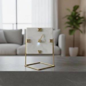 Puede incluir: Un reloj cuadrado de mármol con números romanos y manecillas doradas. El reloj está sobre un soporte de metal dorado. El diseño es minimalista y moderno, adecuado para la decoración del hogar.