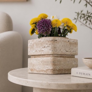 May include: A square beige travertine stone vase with a porous, textured surface and a horizontal recessed groove around the center. The vase is filled with a vibrant bouquet of yellow and purple chrysanthemums and white flowers.