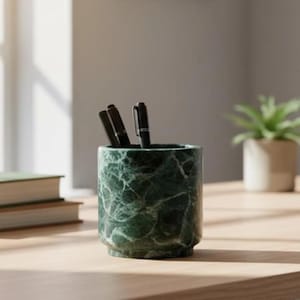 Peut inclure: Un pot à crayons cylindrique en marbre vert foncé avec des veines blanches marquées. L'objet présente une base étagée et contient trois stylos noirs disposés verticalement. La surface du marbre est polie et présente un aspect lisse.