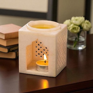 Könnte beinhalten: Ein quadratischer, cremefarbener Marmor-Diffusor für ätherische Öle mit einer Kerze im Inneren. Der Diffusor hat ein dekoratives Ausschnittmuster an den Seiten und eine runde Vertiefung oben. Ein kleiner Strauß weißer Blumen steht in einer Glasvase.