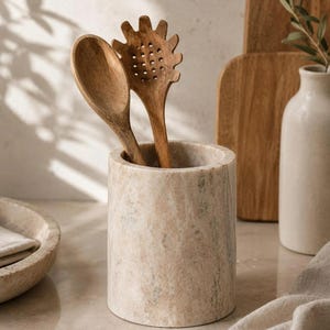 Peut inclure: Un porte-ustensiles en marbre beige avec deux ustensiles de cuisine en bois. Les ustensiles en bois sont une cuillère et une cuillère à spaghetti ajourée. Des planches à découper en bois et un vase sont en arrière-plan.