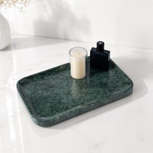 May include: A rectangular dark green marble tray with rounded corners. Resting on the tray is a cylindrical white wax candle in a clear glass holder and a small, square black glass bottle with a black cap, featuring a white logo on the front.