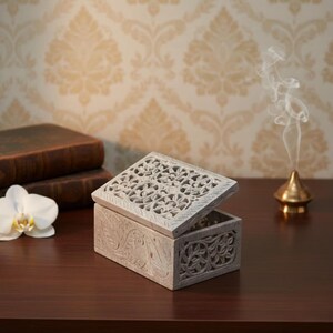 May include: A gray, carved stone box with an open, intricately patterned lid. The box features floral and scroll designs. A white orchid and two old books are on the dark wooden surface. A small gold incense burner emits smoke.