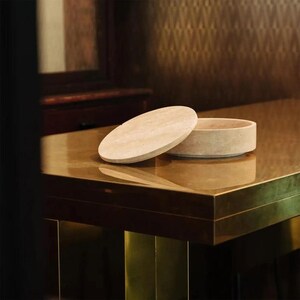 Op de afbeelding: Een beige, cirkelvormige container met een bijpassend deksel, rustend op een reflecterend, goudkleurig oppervlak. De container is gemaakt van een lichtgekleurd steenmateriaal. De achtergrond bevat een donkere houten kast en behang met patronen.