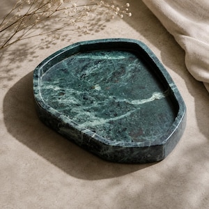 May include: A dark green marble tray with an irregular, organic shape and a smooth, polished surface. The stone features prominent white veining and deep forest green tones throughout the base, with raised edges forming a shallow basin.