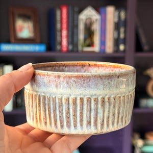 May include: A small, handmade ceramic bowl with a ribbed exterior. The bowl features a blend of colors, including cream, brown, and hints of purple and red. The interior has a glossy finish. The bowl is held in a hand.