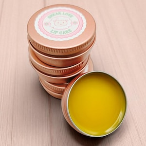 Puede incluir: Una pila de latas de bálsamo labial de color oro rosa, una abierta que revela un bálsamo amarillo. Las tapas tienen una etiqueta rosa y blanca que dice "Shear Love Lip Care" con una ilustración de oso. Las latas de cuidado labial están sobre una superficie de madera rosa claro.