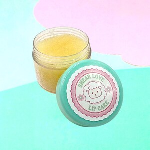 Puede incluir: Un frasco transparente de exfoliante labial amarillo junto a una tapa verde azulado con una etiqueta rosa y blanca. La etiqueta dice "SHEAR LOVE LIP CARE" con una ilustración de oveja de dibujos animados. El fondo es una mezcla de rosa y verde azulado.