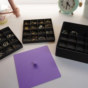 May include: Black jewellery organisers filled with rings, alongside a purple lid. The organisers are divided into square compartments, showcasing rings of various styles and colours. The scene is set on a white surface.