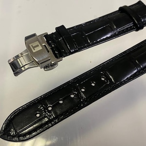 May include: Black leather watch strap with a stainless steel clasp. The strap has a crocodile pattern and is designed for a watch. The clasp features the Tissot logo.