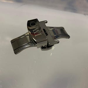 Puede incluir: Un cierre de reloj plateado con las letras "IWC" grabadas. El cierre es de metal y tiene un diseño plegable. El cierre del reloj está diseñado para asegurar una correa de reloj.