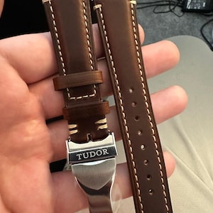 Puede incluir: Correa de reloj de cuero marrón con costuras color crema. La correa presenta una hebilla plateada con el nombre de la marca "TUDOR" grabado. La correa está diseñada para un reloj y se muestra en primer plano.