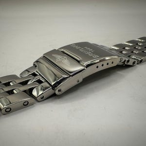 Könnte beinhalten: Nahaufnahme eines Edelstahl-Uhrenarmbands mit Faltschließe. Auf der Schließe steht "BREITLING 1884" und ein Logo. Das Armband besteht aus ineinandergreifenden Gliedern.