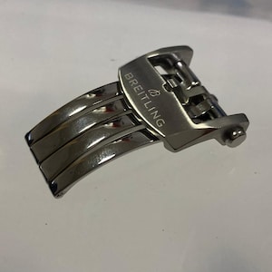 May include: A silver-toned watch clasp with the brand name "BREITLING" engraved on the top. The clasp is made of metal and has a folding design with multiple articulated segments.