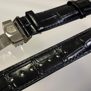 Peut inclure: Bracelet de montre noir avec un motif crocodile. Le bracelet est doté d'un fermoir argenté avec le logo "TAG Heuer". Le bracelet est fait d'un matériau brillant et texturé.