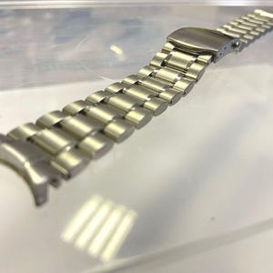 May include: A silver-toned watch band with a brushed metal finish. The band is composed of multiple rectangular links connected by smaller, rounded links. The clasp is a fold-over style. The band is designed to attach to a watch case.