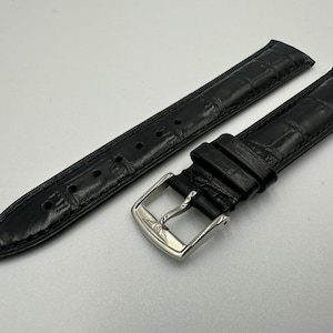May include: Black leather watch strap with a crocodile pattern. The strap has a silver-coloured buckle. The watch strap is new and unused.