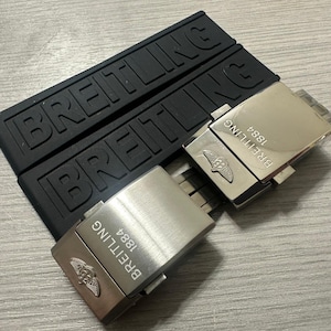 Puede incluir: Correas de reloj de goma negra y dos cierres de reloj plateados. Las correas est&aacute;n grabadas con la palabra "BREITLING". Los cierres est&aacute;n grabados con "BREITLING 1884" y un logotipo.