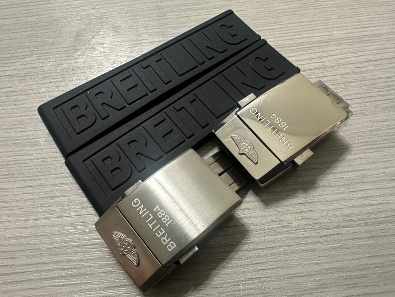 Puede incluir: Correas de reloj de goma negra y dos cierres de reloj plateados. Las correas est&aacute;n grabadas con la palabra "BREITLING". Los cierres est&aacute;n grabados con "BREITLING 1884" y un logotipo.
