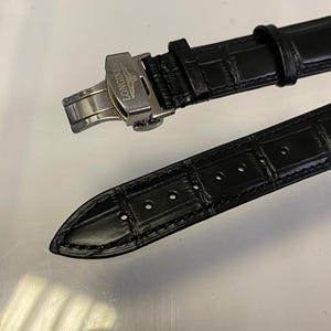 May include: Black leather watch strap with a stainless steel clasp. The strap has a textured, crocodile-like pattern. The clasp is marked with the brand name "Longines". The strap is in two pieces, with one piece having several holes.