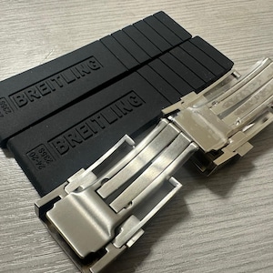 Puede incluir: Correas de reloj de goma negra con el nombre de la marca "Breitling" en relieve, junto con un cierre de reloj plateado. Las correas muestran la informaci&oacute;n de tama&ntilde;o "24-20 235S".