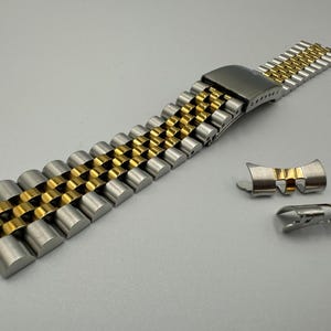 May include: A two-tone stainless steel and gold watch strap with a folding clasp. The strap features alternating gold and silver links. Two additional strap pieces are also included.
