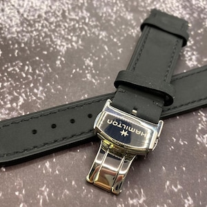 Peut inclure: Un bracelet de montre noir doté d'une boucle déployante en métal argenté. La boucle est gravée du mot Hamilton et d'un petit logo. Le bracelet présente des coutures visibles sur les bords et plusieurs trous de réglage pour un ajustement sûr.