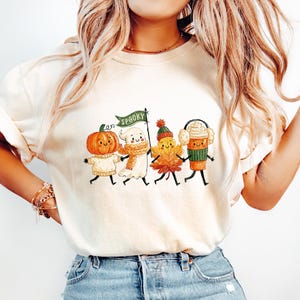 May include: A cream-colored short-sleeve t-shirt featuring a graphic of four cartoon characters: a pumpkin in a sweater, a ghost with a scarf holding a green flag that says SPOOKY, a leaf wearing a beanie, and a coffee cup with earmuffs.