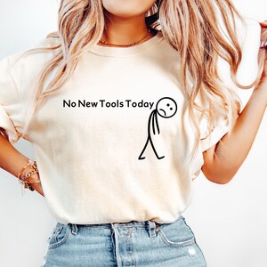 May include: Cream-colored t-shirt with the text "No New Tools Today" and a dejected stick figure graphic. The shirt is paired with light blue denim jeans.