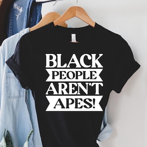 May include: A black t-shirt with the white text "BLACK PEOPLE AREN'T APES!". The shirt is hanging on a wooden hanger with other clothing items in the background.