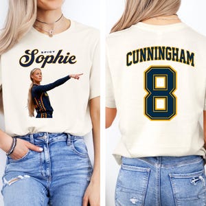 May include: A cream-colored t-shirt featuring a graphic on the front with the text Spicy Sophie and an image of a basketball player pointing. The back of the shirt displays the name CUNNINGHAM in dark blue letters above the large dark blue number 8.