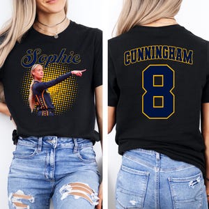 May include: A black t-shirt featuring a graphic of a basketball player pointing, with the name Sophie in blue and yellow script above. The back of the shirt displays the name CUNNINGHAM in blue and yellow block letters above the large number 8.