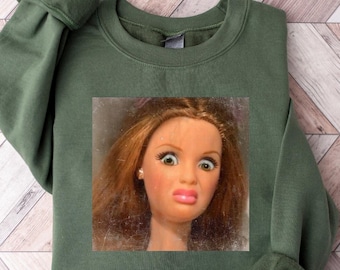 May include: Military green sweatshirt featuring a doll face graphic. The doll has wide eyes and a pursed lip. The sweatshirt is laid flat on a wooden surface.