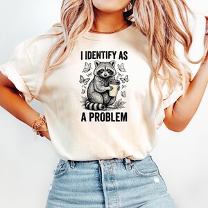 May include: A cream-colored t-shirt featuring a black ink-style graphic of a raccoon holding a coffee cup, surrounded by butterflies. The text above the raccoon reads I IDENTIFY AS and the text below the raccoon reads A PROBLEM in bold letters.