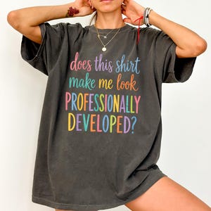 May include: A charcoal gray short-sleeve t-shirt featuring the colorful text "does this shirt make me look PROFESSIONALLY DEVELOPED?" printed on the front. The shirt has a crew neckline and a loose, relaxed fit with a washed-out fabric appearance.