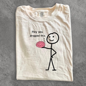 May include: A cream-colored t-shirt featuring a black stick figure graphic holding a pink brain. The text above the brain reads, "Hey you dropped this." The shirt has a Comfort Colors brand label visible at the inner neckline, printed on cotton fabric.
