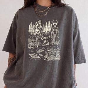 May include: A charcoal gray t-shirt featuring a cream-colored graphic print of Bigfoot, the Loch Ness Monster, and several aliens with flying saucers. The text "Respect the local" is printed in the center of the design in a cursive font.