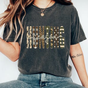 May include: A charcoal gray short-sleeve t-shirt featuring the word HUNTING in block letters with varied patterns including deer silhouettes, plaid, leopard print, stars, camouflage, and polka dots, with the word season in white script.