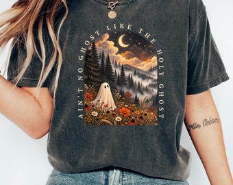 May include: A charcoal gray t-shirt featuring a graphic print of a ghost in a field of wildflowers, pine trees, and a crescent moon under a cloudy sky. The text "AIN'T NO GHOST LIKE THE HOLY GHOST" is arched over the illustration in a serif font.